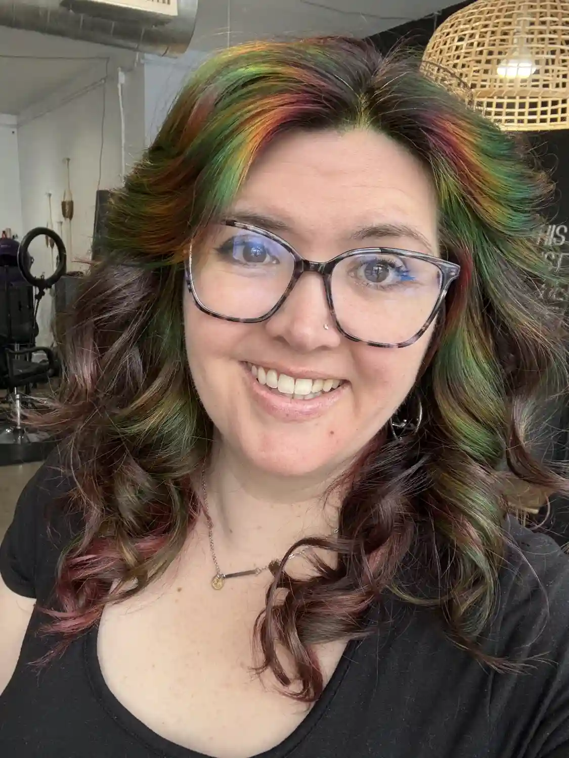 Smiling master stylist Kelly at Studio 303, wearing glasses and showcasing shoulder-length curled hair with an oil spill color blend of green and red tones on a dark base, inside the salon.