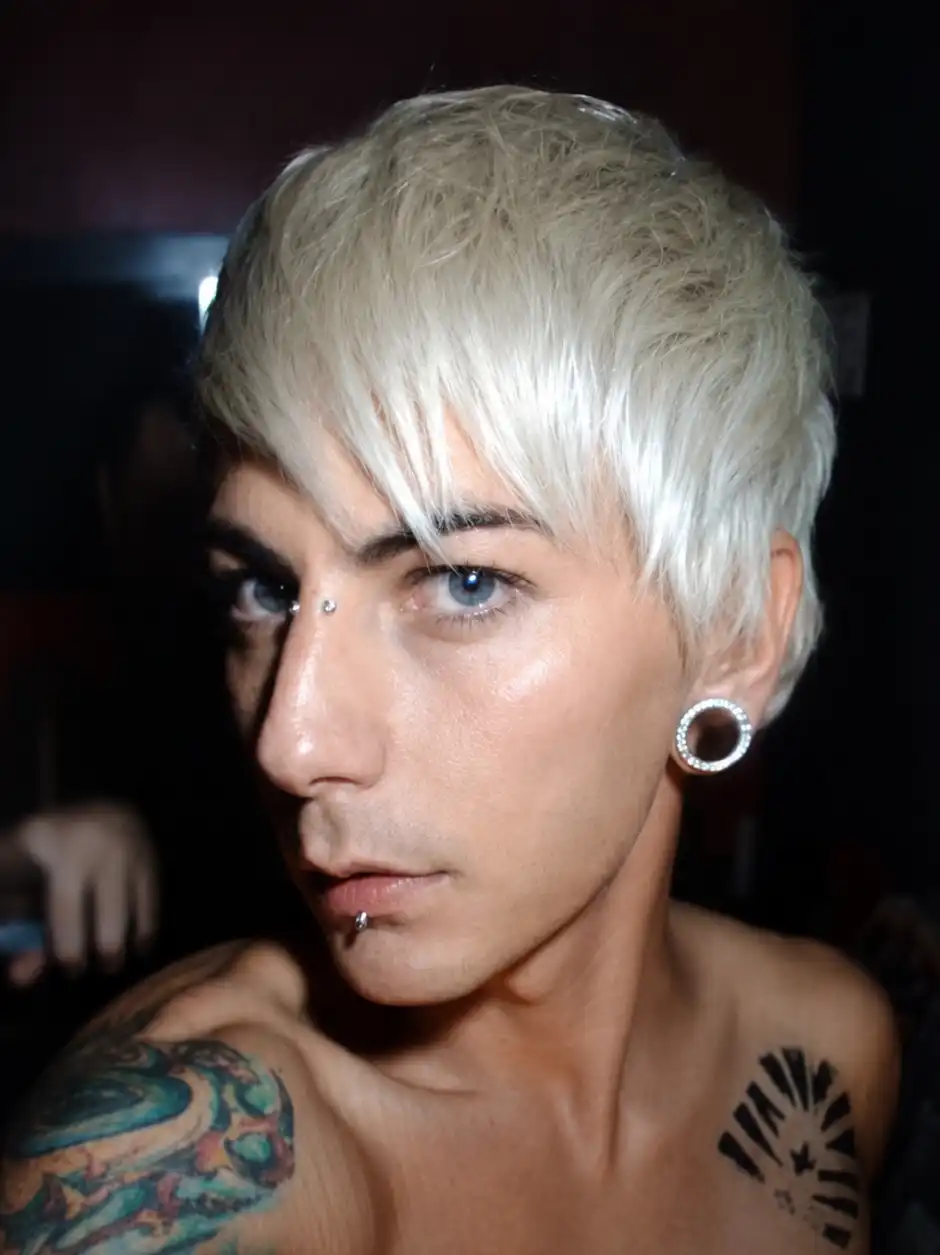 Person with platinum blonde short hair, facial piercings, and tattoos posing in dark dramatic lighting.