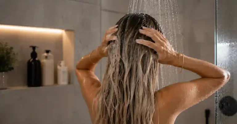 Woman washing long blonde hair extensions in shower, water rinsing downward from scalp to ends