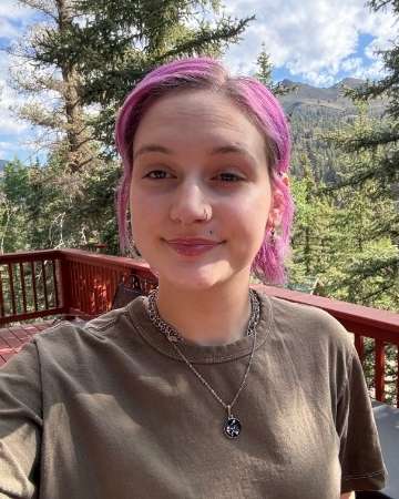 Taryn, hairstylist at Studio 303 Salon, smiling for a selfie on an outdoor wooden deck with trees and mountains in the background.